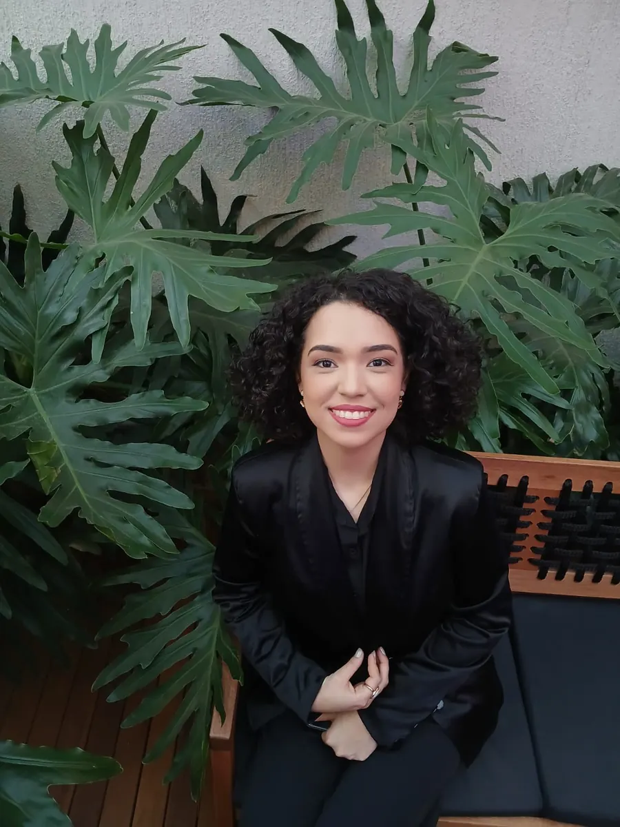 Rebeca Costa Ferreira, psicóloga, sorrindo em ambiente com folhagem.